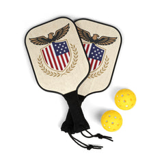 Two wooden pickleball paddles with black grip tape and a patriotic shield graphic, alongside two yellow perforated pickleball balls.